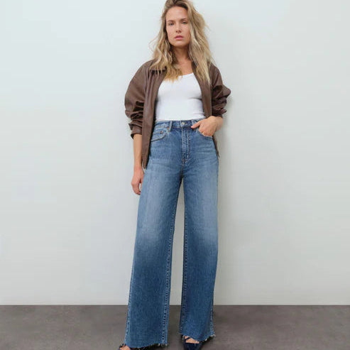Woman wearing a brown jacket, white top, and blue jeans against a plain background