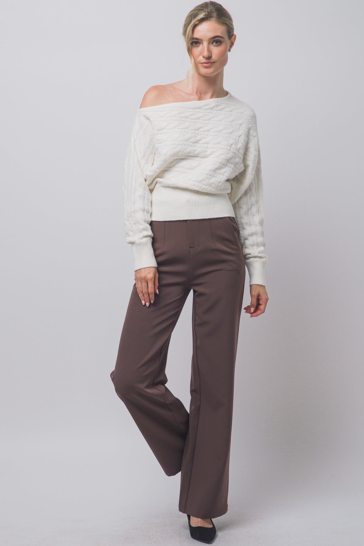 Ivory cable-knit off-shoulder sweater styled with brown trousers