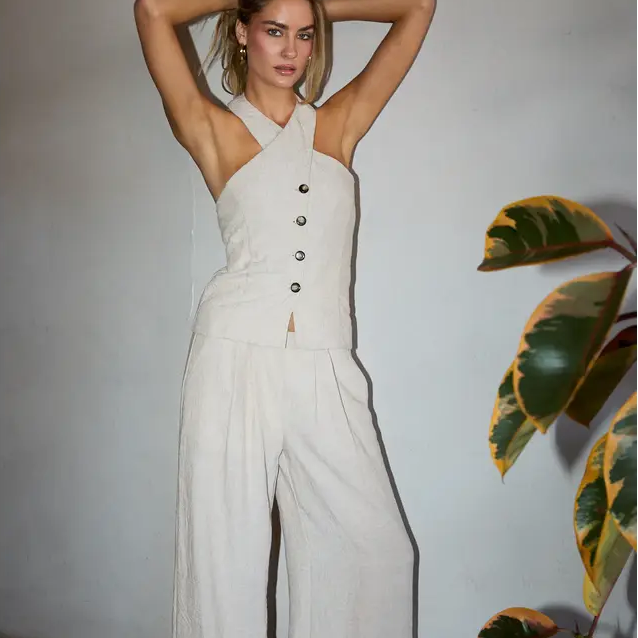Woman wearing a white jumpsuit with buttons, standing against a plain wall with a plant to the side.