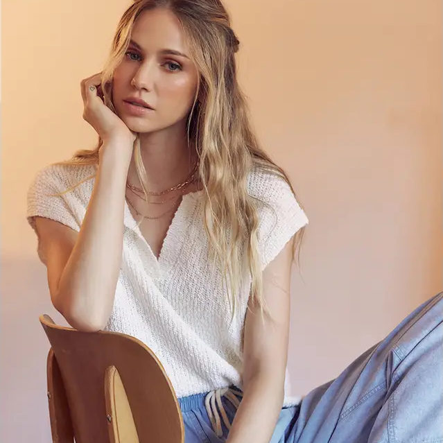 Woman sitting on a wooden chair wearing a white top and light blue pants against a beige wall.